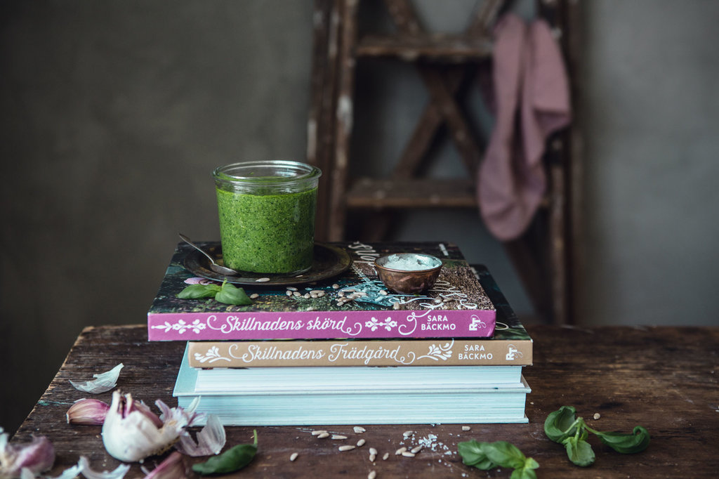 En härligt, grön pesto står i en glasburk på böckerna Skillnadens Trädgård och Skillnadens Skörd.