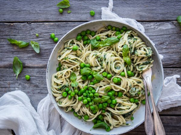 På ett grått träbord står en skål med spagetti med krämig grön sås. Knallgröna ärtor ligger i skålen och runtomkring.