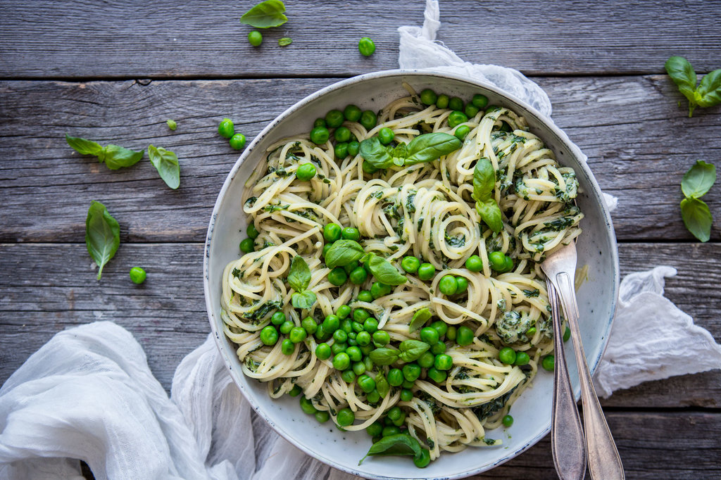 På ett grått träbord står en skål med spagetti med krämig grön sås. Knallgröna ärtor ligger i skålen och runtomkring.