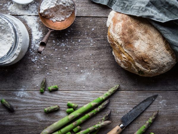 Ett rustikt bröd ligger på ett träbord tillsammans med sparris och mjöl.