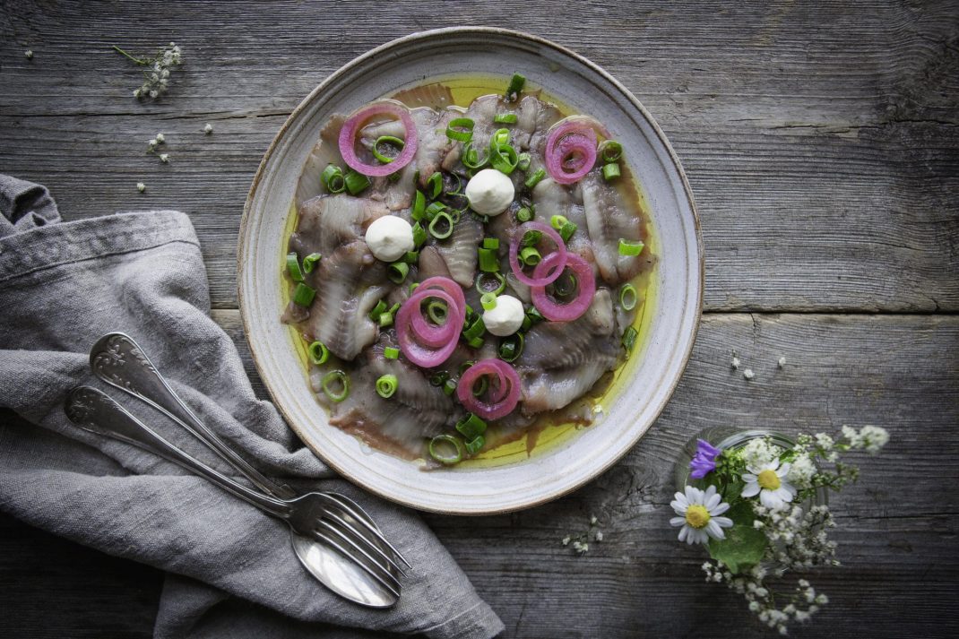 På ett grått träbord står en rustik grå tallrik med tunt skivade matjesfileer, gräslök, picklad rödlök. Bredvid står en liten bukett med midsommarblommor.