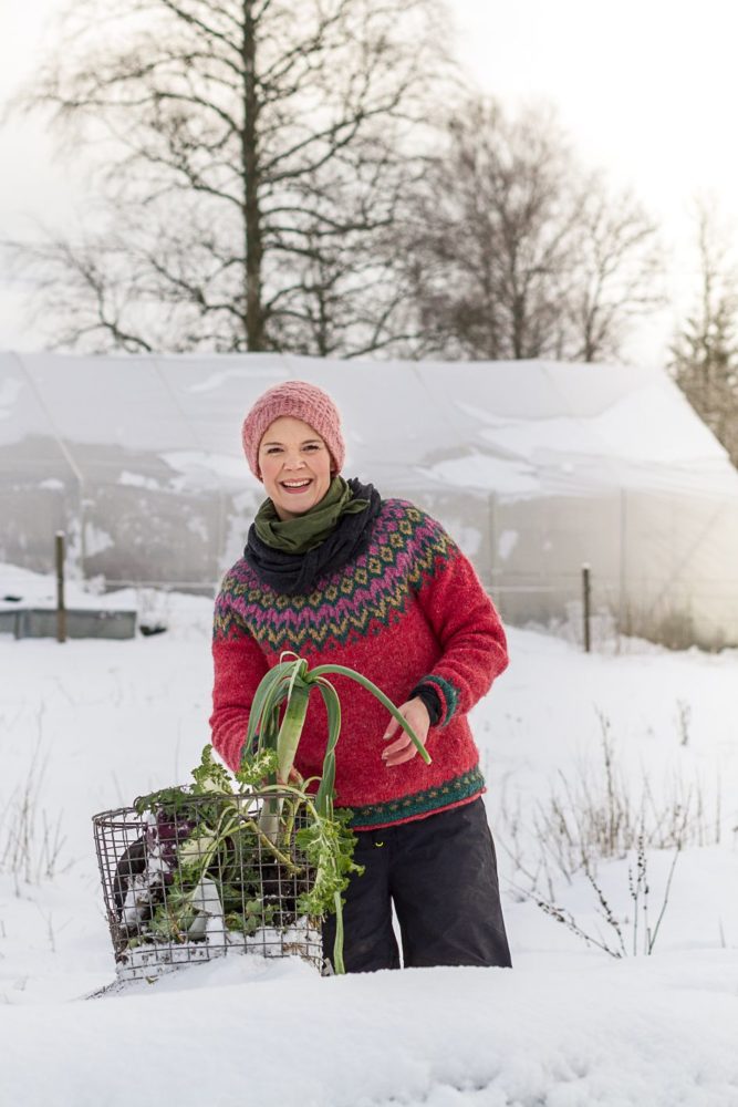 Sara Bäckmo står i en snöig trädgård med en korg ed grönsaker. 