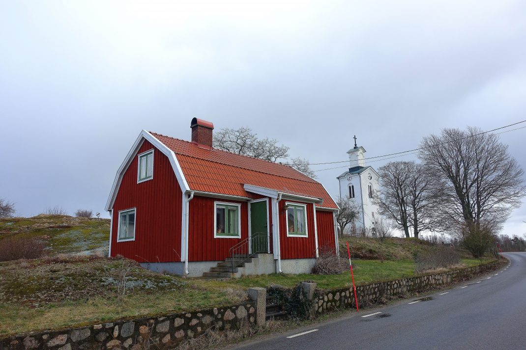 Ett litet rött hus med stenmur framför och en vit kyrka i bakgrunden. 