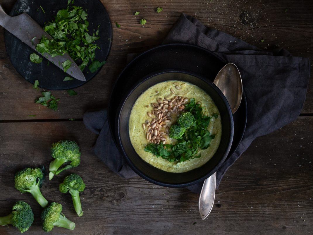 Broccolisoppa i en svart soppskål. Vid sidan syns hackad persilja och broccolibuketter.